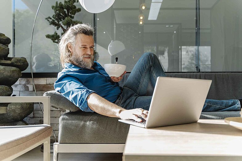 Ein Mann mit langen Haaren und Bart sitzt entspannt auf einem Sofa, hält eine Schüssel in der Hand und bedient mit der anderen ein Laptop auf dem Tisch vor ihm.