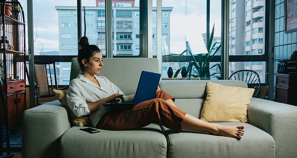 Frau liegt entspannt mit Laptop auf einem grauen Sofa. Im Hintergrund eine Fensterfront
