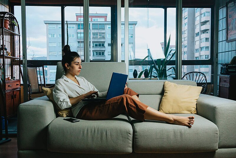 Frau liegt entspannt mit Laptop auf einem grauen Sofa. Im Hintergrund eine Fensterfront