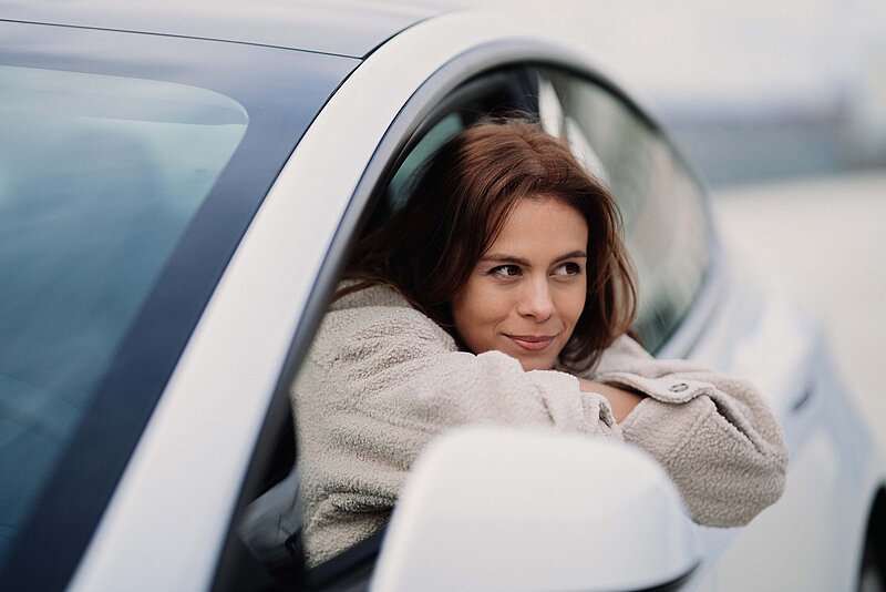 Elektrofahrzeug-jenny-ueberberg-unsplash. Frau schaut aus Fenster von weißem Auto und hat die Arme auf der Autotür abgelegt