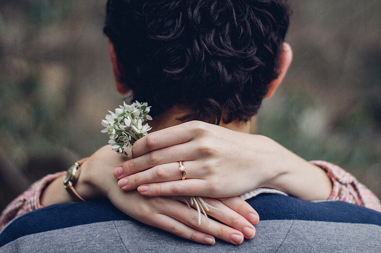 Verheiratete Steuerzahler-Joshua Rodriguez-Unsplash Schultern und Kopf einer Person von hinten. Hände einer zweiten Person mit Ring im Fokus und kleinem Blumenstrauß in der Handim Nachen gefaltet mi