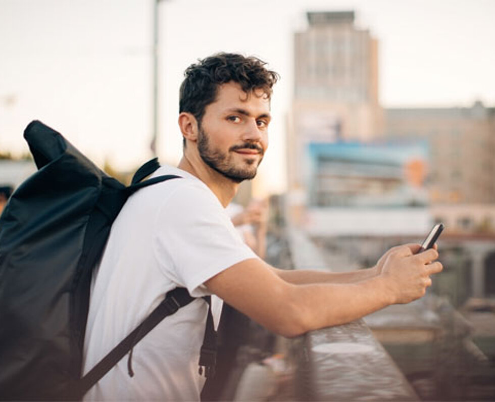 Junger Mann mit Rucksack in Abendstimmung, an einem Geländer stehend das Handy in der Hand, im Hintergrund Hochhäuser.