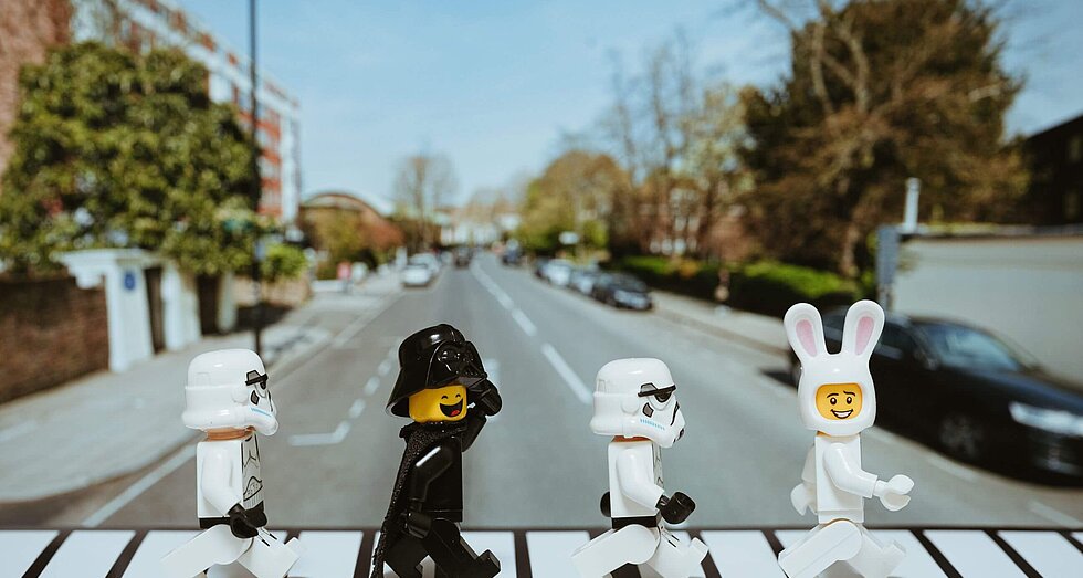 Vier Playmobil Figuren in schwarz und weiß laufen wie die Beatles über einen Zebrastreifen