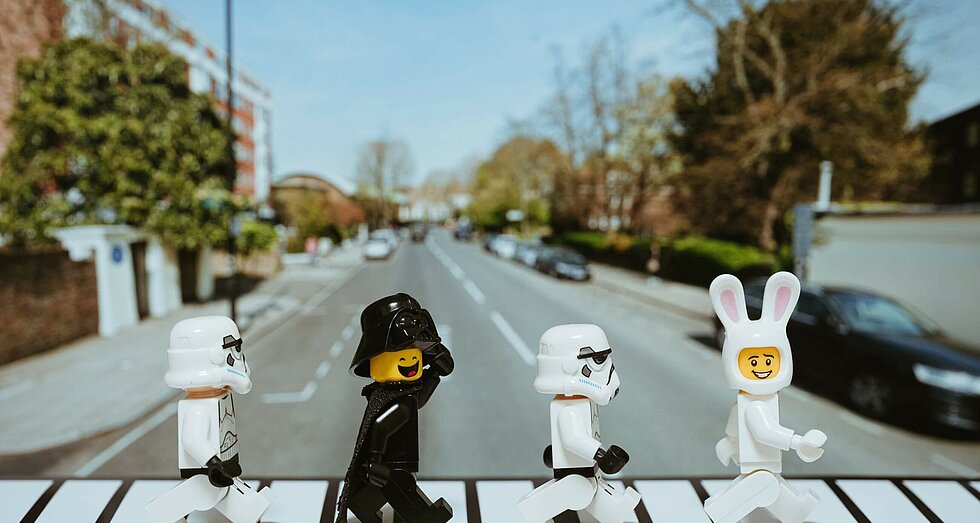 Vier Playmobil Figuren in schwarz und weiß laufen wie die Beatles über einen Zebrastreifen