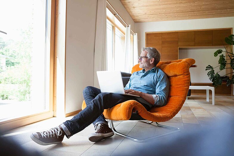 Ein Mann mit Brille sitzt entspannt in einem orangefarbenen Sessel in einem modernen Wohnzimmer, arbeitet an einem Laptop auf seinem Schoß und blickt dabei aus dem Fenster.