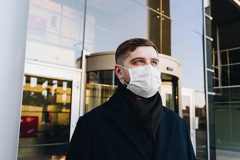 Corona Verlustrücktrag-Anastasiia Chepinska-Unsplash. Man mit Atemschutzmaske vor verspiegelter Gebäudefront