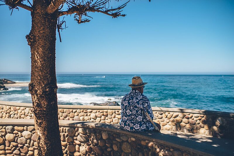 Riester-Rente-Ian Schneider-Unsplash. Mann sitzt auf einer Steinmauer und schaut aufs Meer