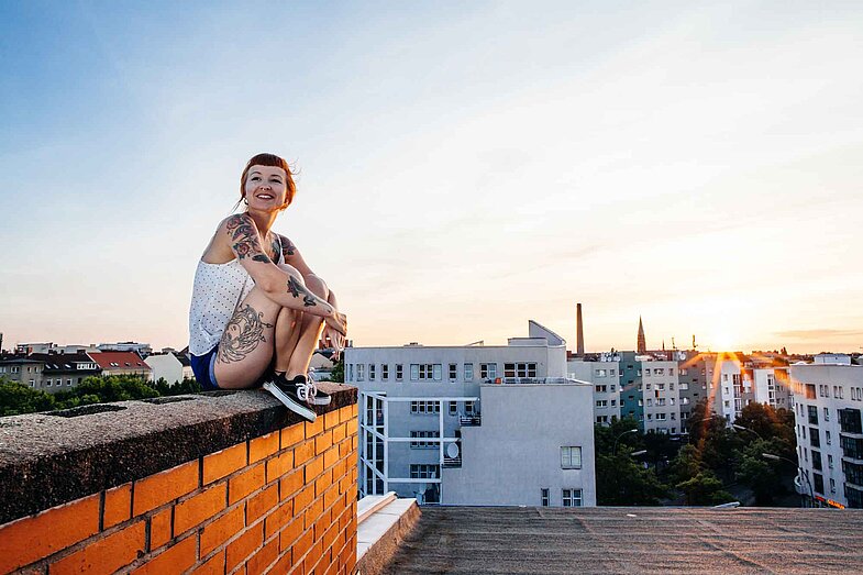 Eine junge Frau mit Tätowierungen sitzt lächelnd auf einer Backsteinmauer am Rand eines Daches