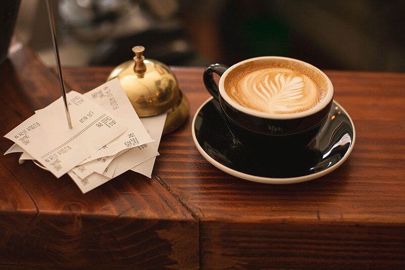 Bonpflicht-Carli Jeen-Unsplash Kaffeetasse auf Holztheke neben einem Stapel Kassenbons