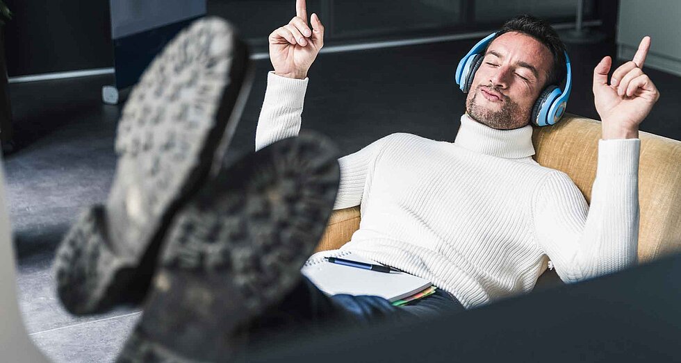 Ein Mann in einem weißen Rollkragenpullover sitzt entspannt mit Kopfhörern in einem Sessel, hat die Füße hochgelegt, die Augen geschlossen und hebt beide Zeigefinger im Takt der Musik.