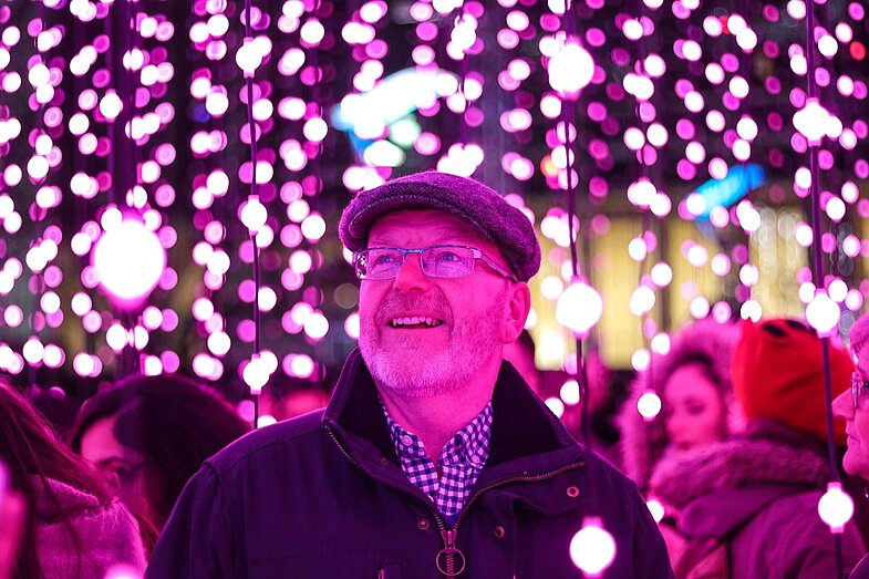 älterer Mann steht vor Lichterketten, Bild ist leicht rosa eingefärbt