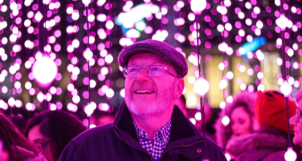 älterer Mann steht vor Lichterketten, Bild ist leicht rosa eingefärbt