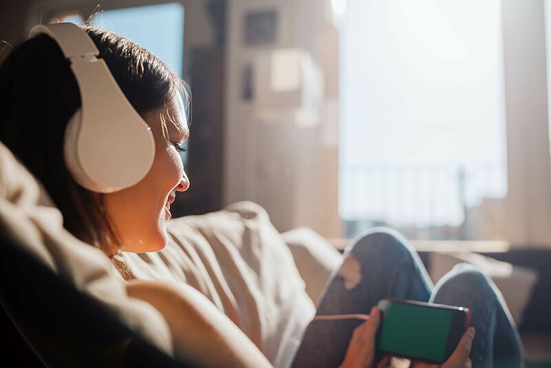 Inflationsausgleichpraemie-Guido-Mieth-Gettyimage Junge musikhörende Frau auf Sofa in sonnendurchflutetem Raum