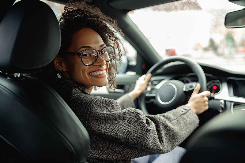 Eine Frau mit Brille sitzt lächelnd am Steuer eines Autos und schaut über die Schulter nach hinten.